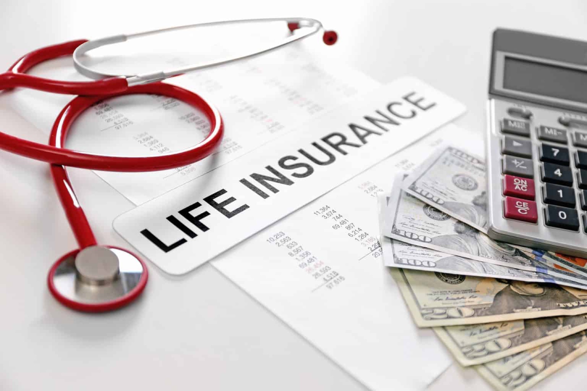 Pile of money sitting under a calculator on a table next to a red stethoscope and a sign reading "life insurance."