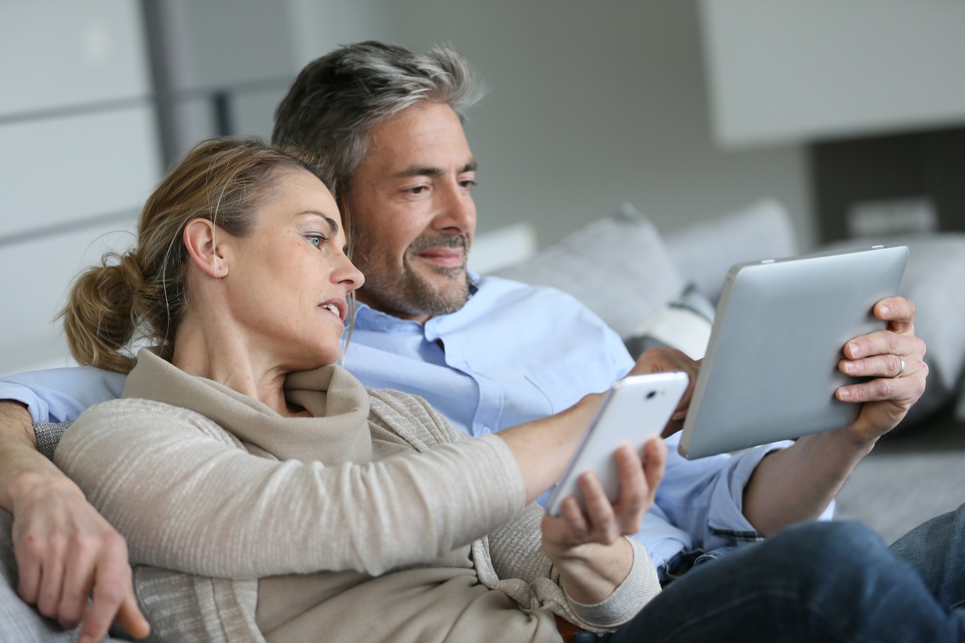 A middle-aged couple is sitting next to each other on the couch, using a smartphone and tablet to review their retirement plan.